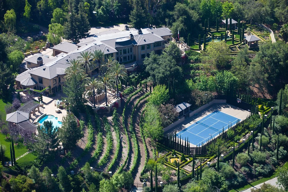 A large estate with a mansion, a swimming pool, a tennis court, and lush gardens surrounded by trees.
