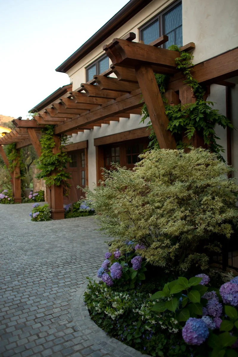 A house with a wooden pergola, green climbing plants, and colorful flowers in a garden bed, paved walkway in front.