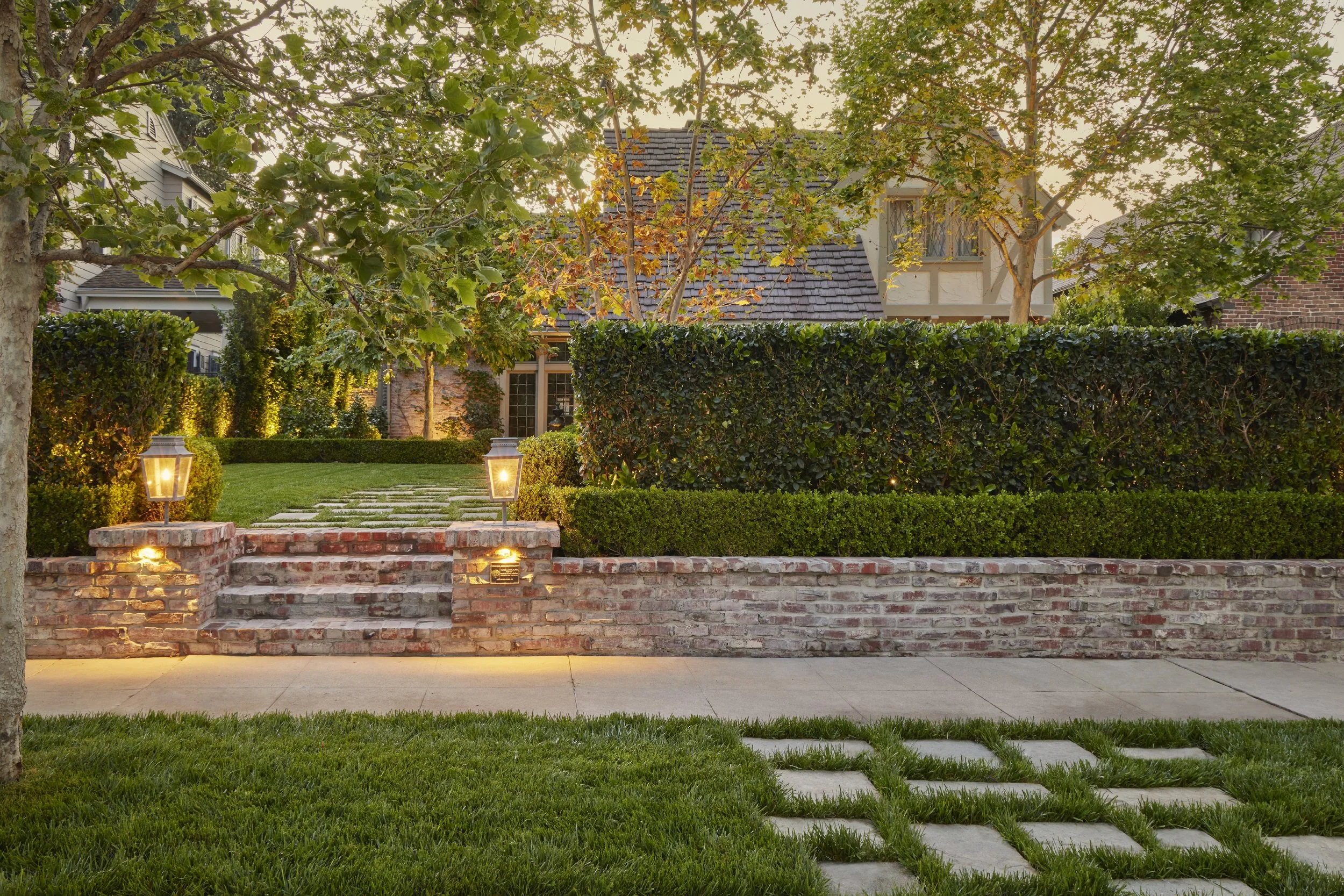 A front yard with a brick wall, stone steps, green grass, trees, and decorative lanterns near the steps, with a house visible behind the trees.