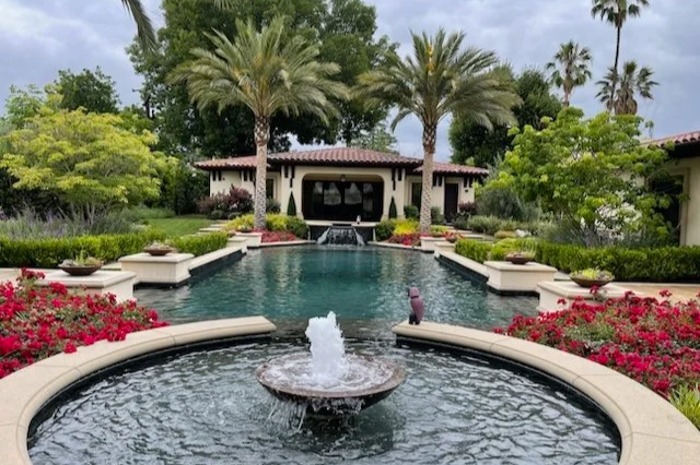 Luxury backyard with a rectangular swimming pool, surrounded by palm trees, lush greenery, and colorful flowers, with a small fountain in the foreground.