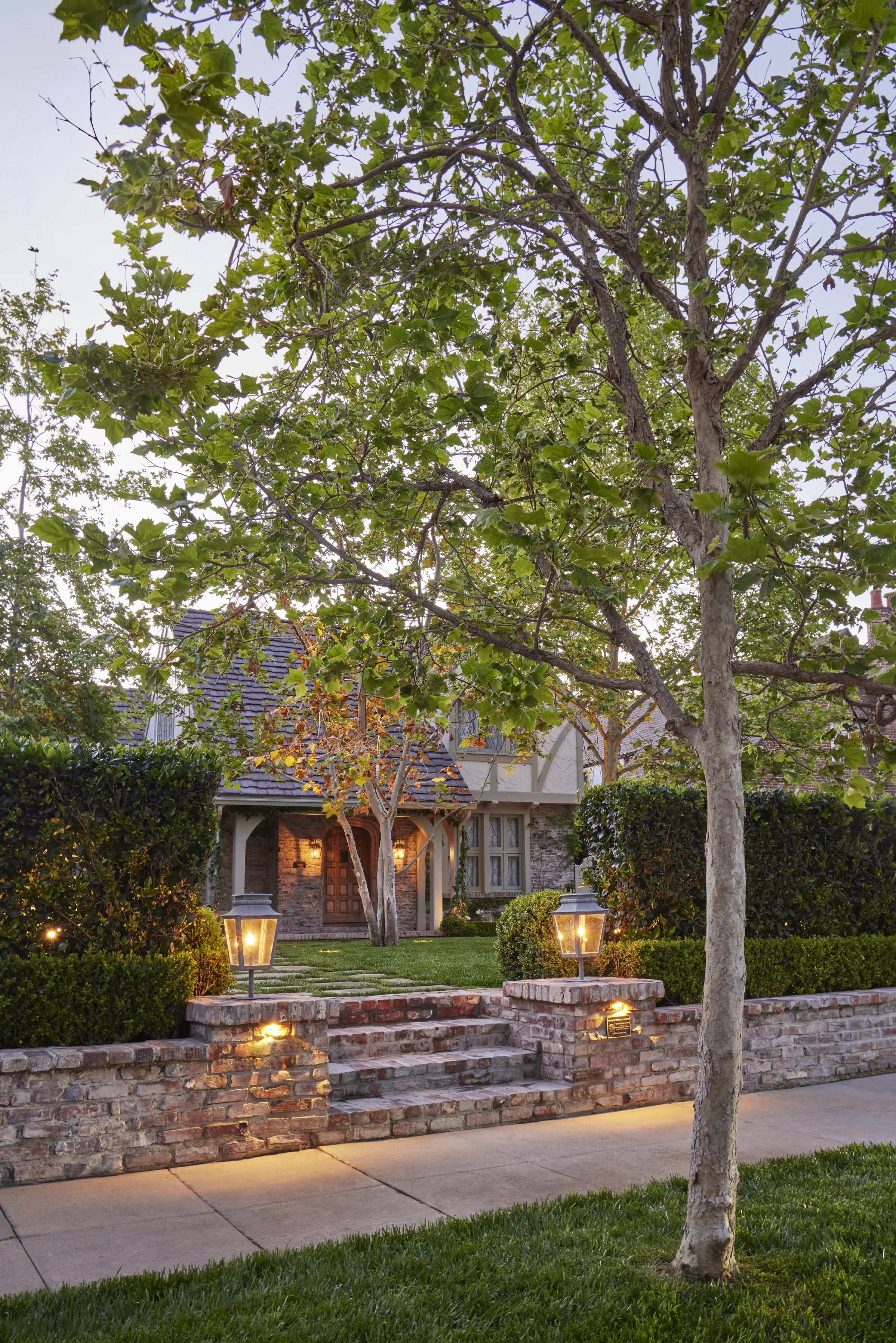Front view of a house with brick steps, brick front wall, a wooden door, and two lanterns lighting the walkway. Two trees with green leaves are in the foreground, and a well-maintained lawn and shrubs surround the house. The setup appears to be during dusk.