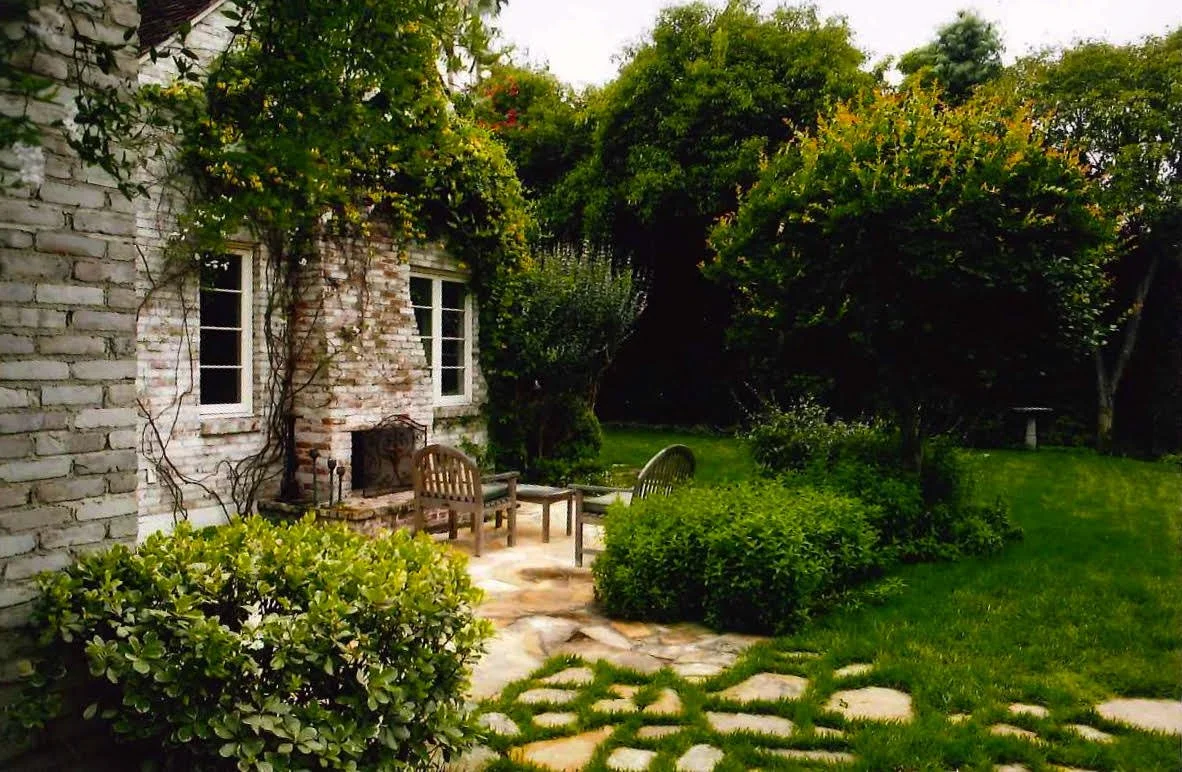 A cozy outdoor garden with a brick house wall, two small windows, and a brick fireplace. There are garden chairs, lush bushes, trees, and a green lawn with stepping stones.