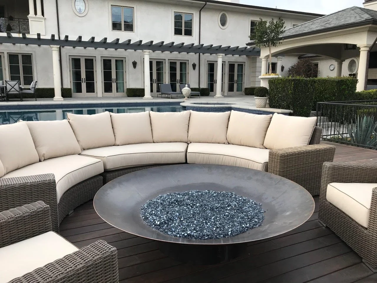 Outdoor patio with a large curved cream-colored cushioned sectional sofa surrounding a fire pit table with blue glass chips, overlooking a swimming pool and a large house in the background.