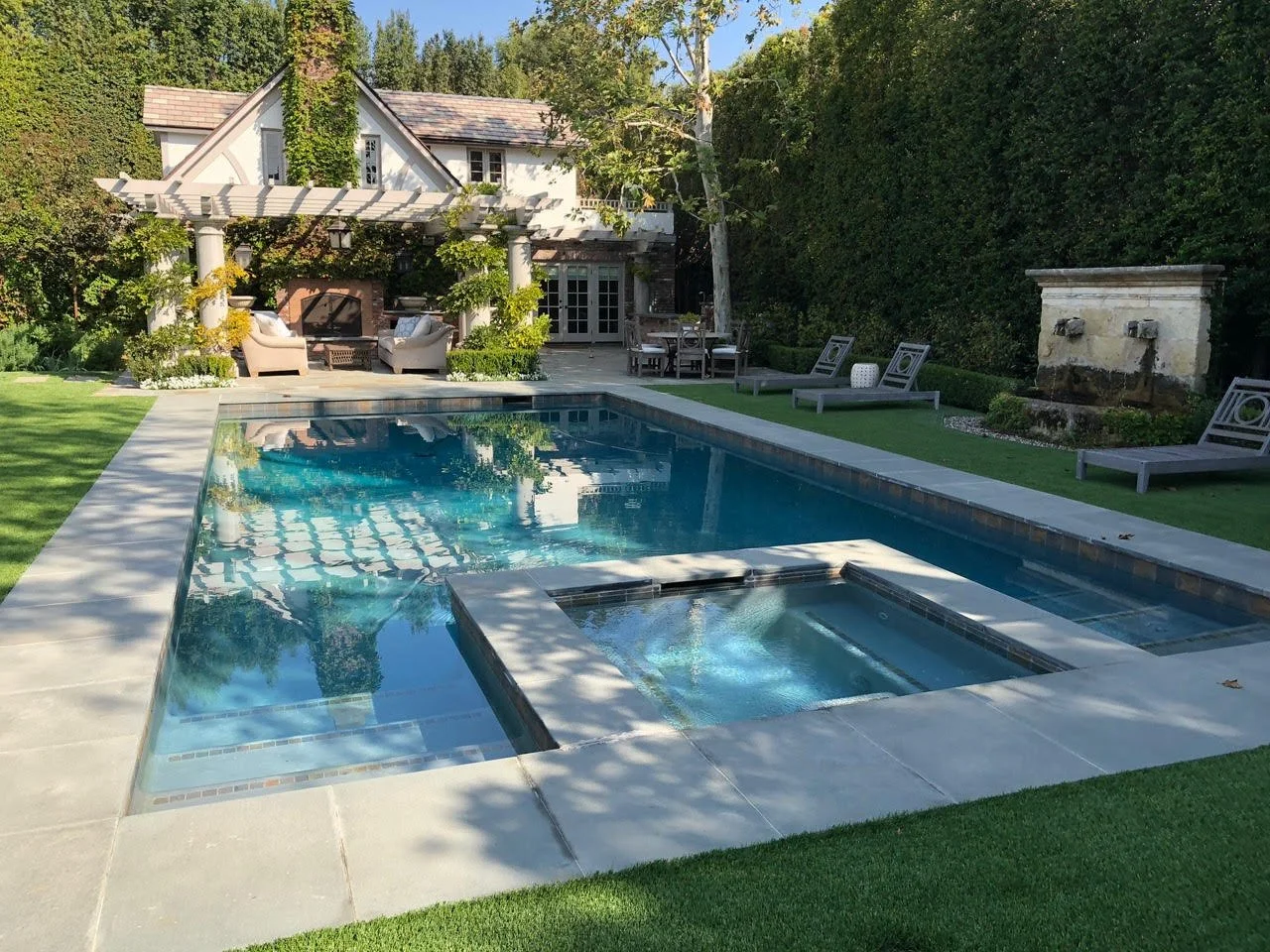 Backyard with a rectangular swimming pool, lounge chairs, outdoor seating, and a house with a pergola and lush greenery.