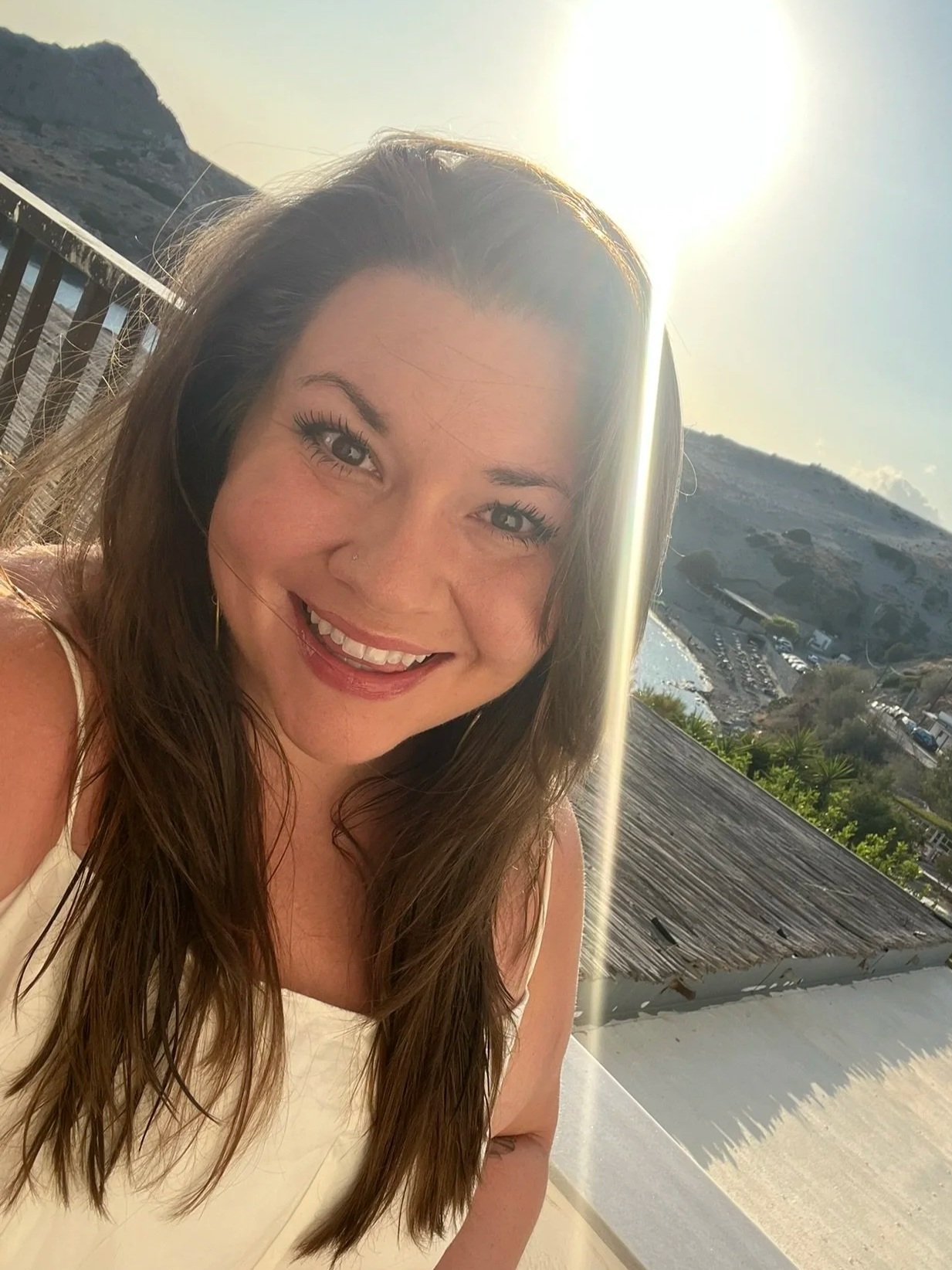 A woman with long brown hair, smiling with a nose piercing, in front of a sunny outdoor landscape with mountains and water.