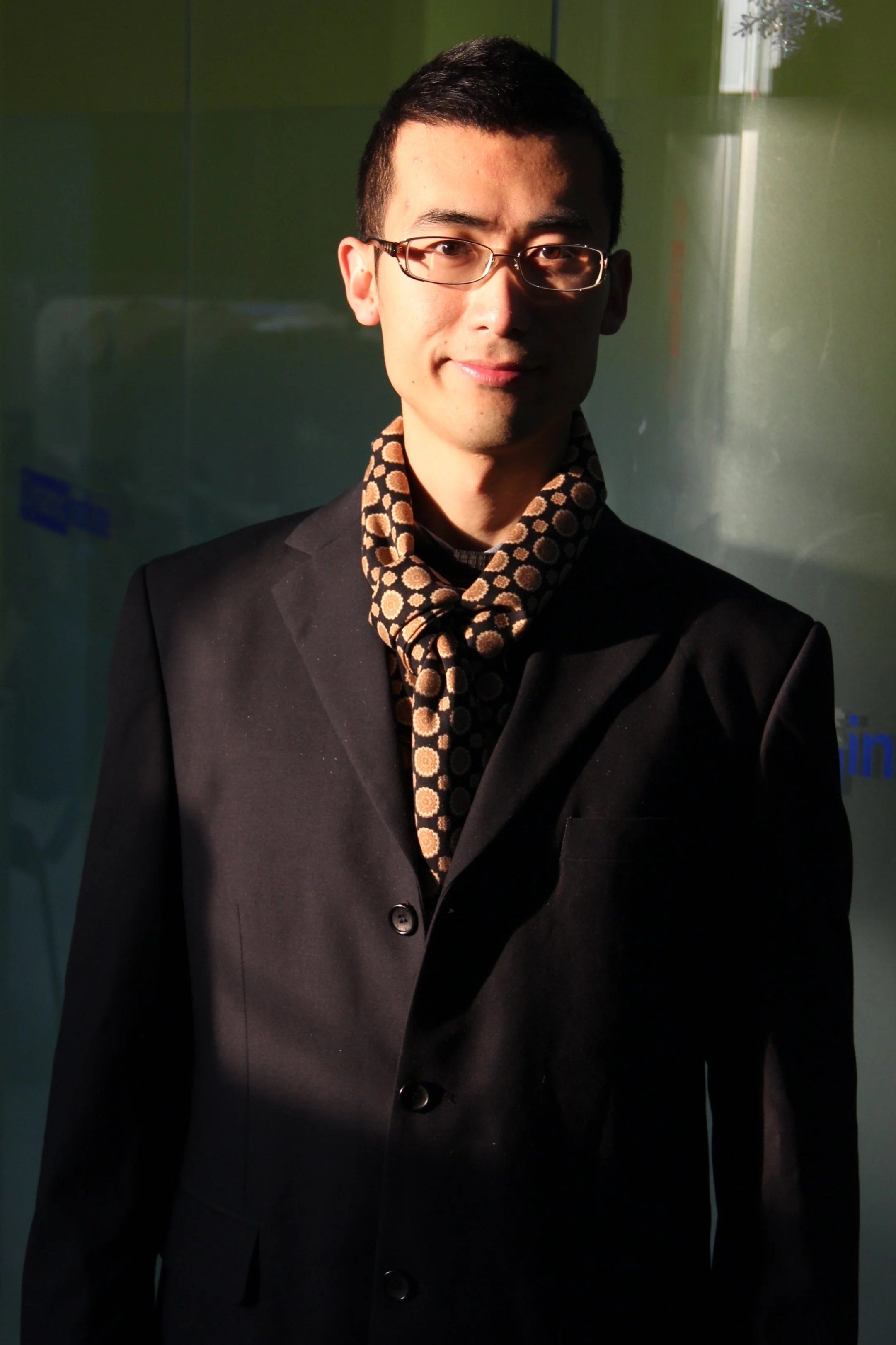 A young man in glasses wearing a black suit and a patterned scarf, standing in front of a slightly reflective wall with partial text, illuminated by a bright light.
