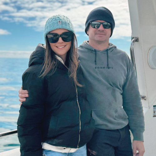 A smiling woman and a man standing together on a boat, dressed in winter clothing with sunglasses, with the ocean and sky in the background.