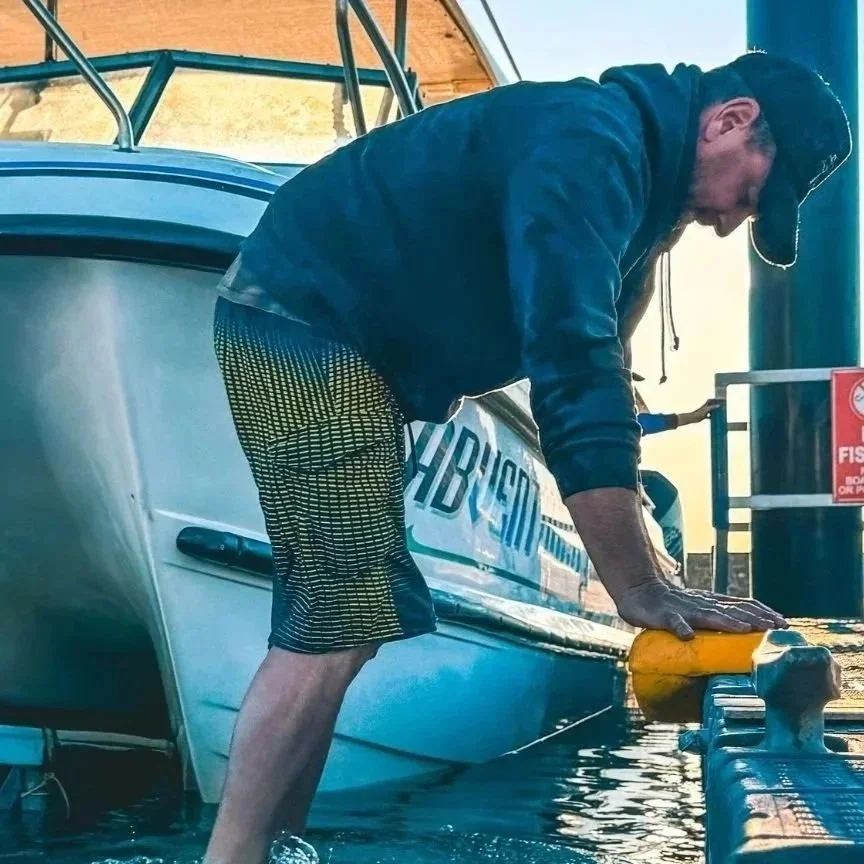 Man leaning over the edge of a boat, holding a yellow fender, docked at a marina during daytime.