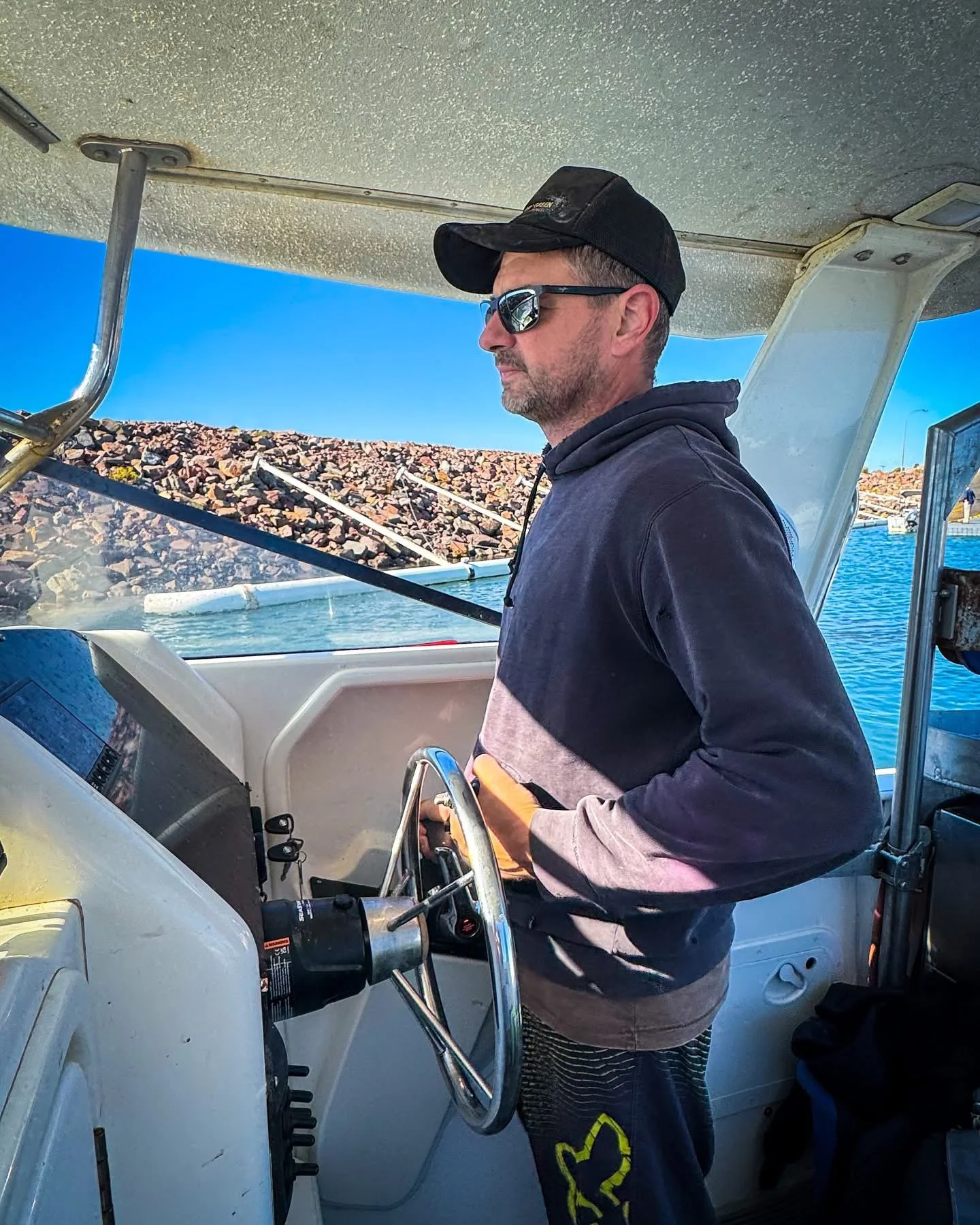 Man in sunglasses and black cap steering a boat, background of blue sky and rocky shoreline.