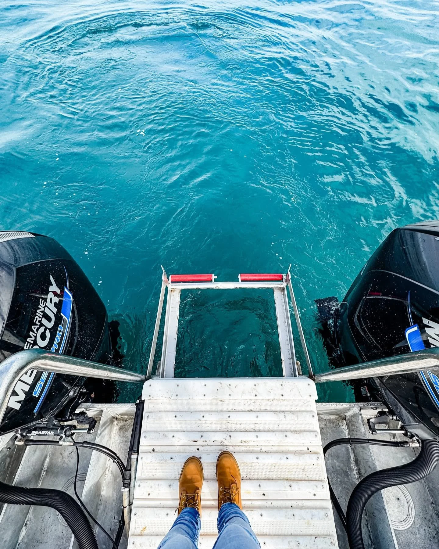 View from a boat's watercraft showing two outboard motors, a metal dock ladder, and a person's legs in yellow shoes and blue jeans standing on the boat's platform over blue water.