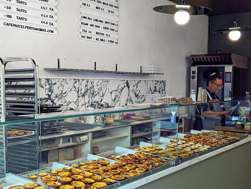 Display case filled with various baked pastries in a cafe. Behind the counter, a woman is working, with a menu board above and a marble backsplash.