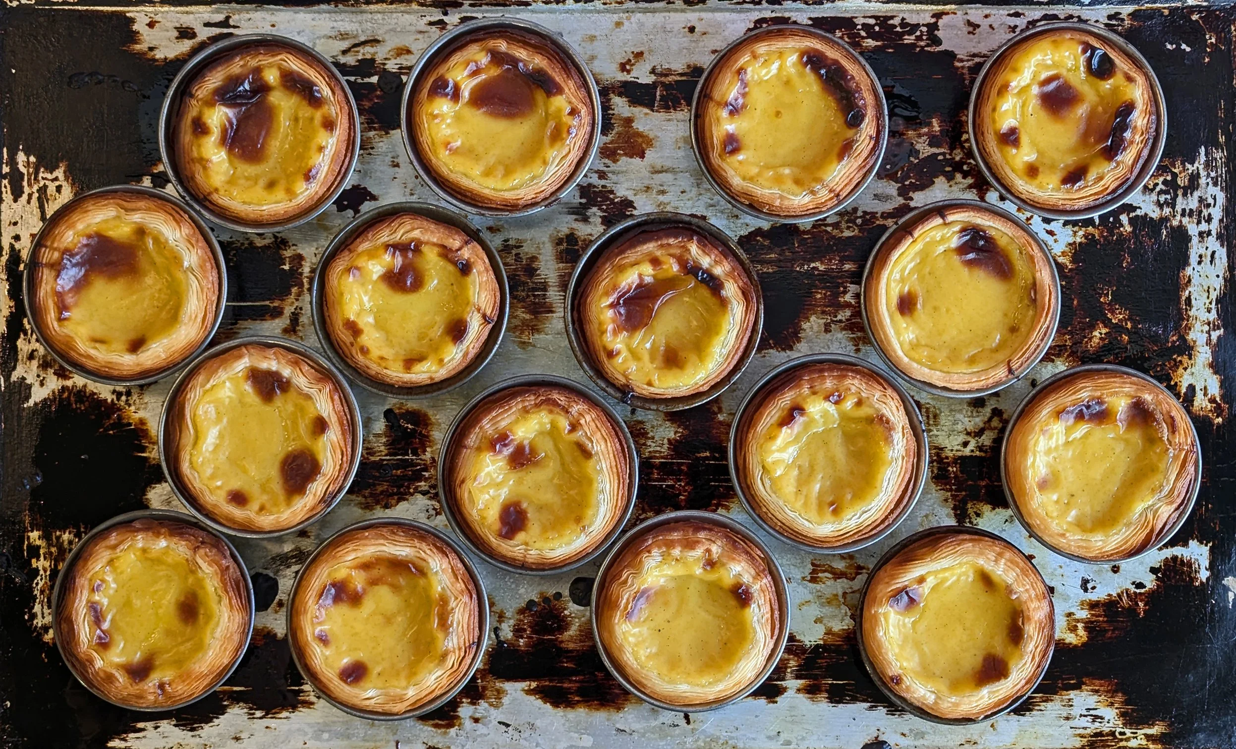 A group of 17 Portuguese egg tarts arranged on a rustic baking sheet.