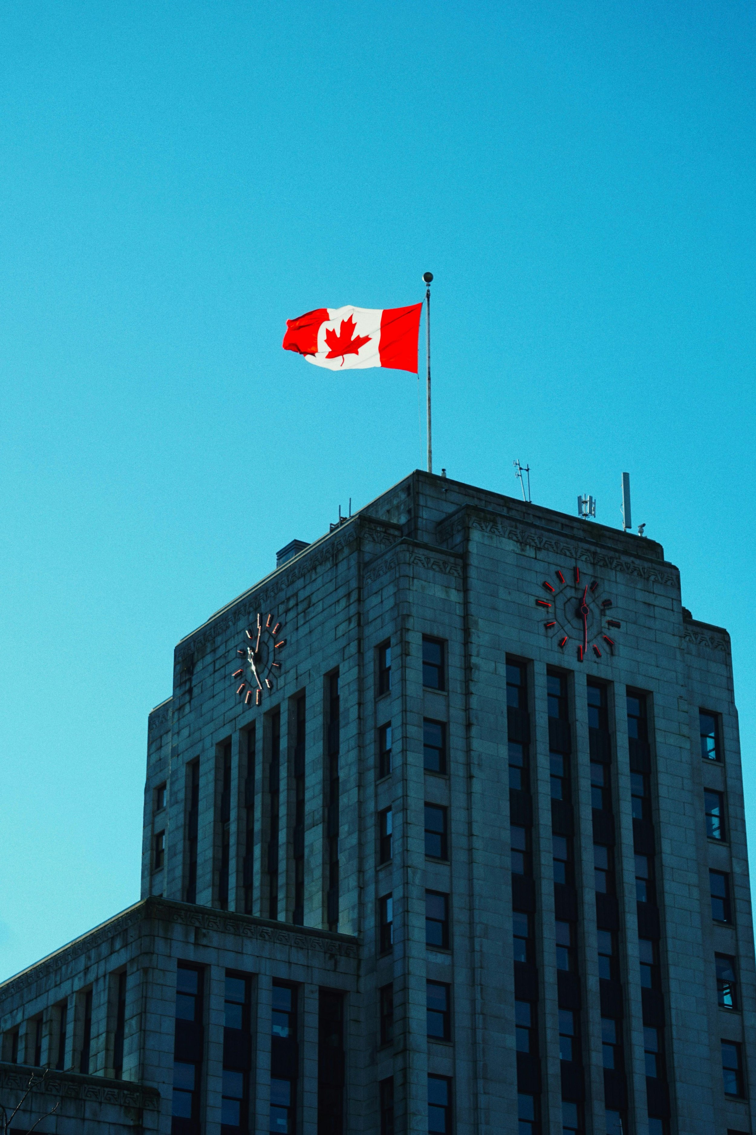 A tall building with a clock on each side and the Canadian flag flying on top against a clear blue sky.