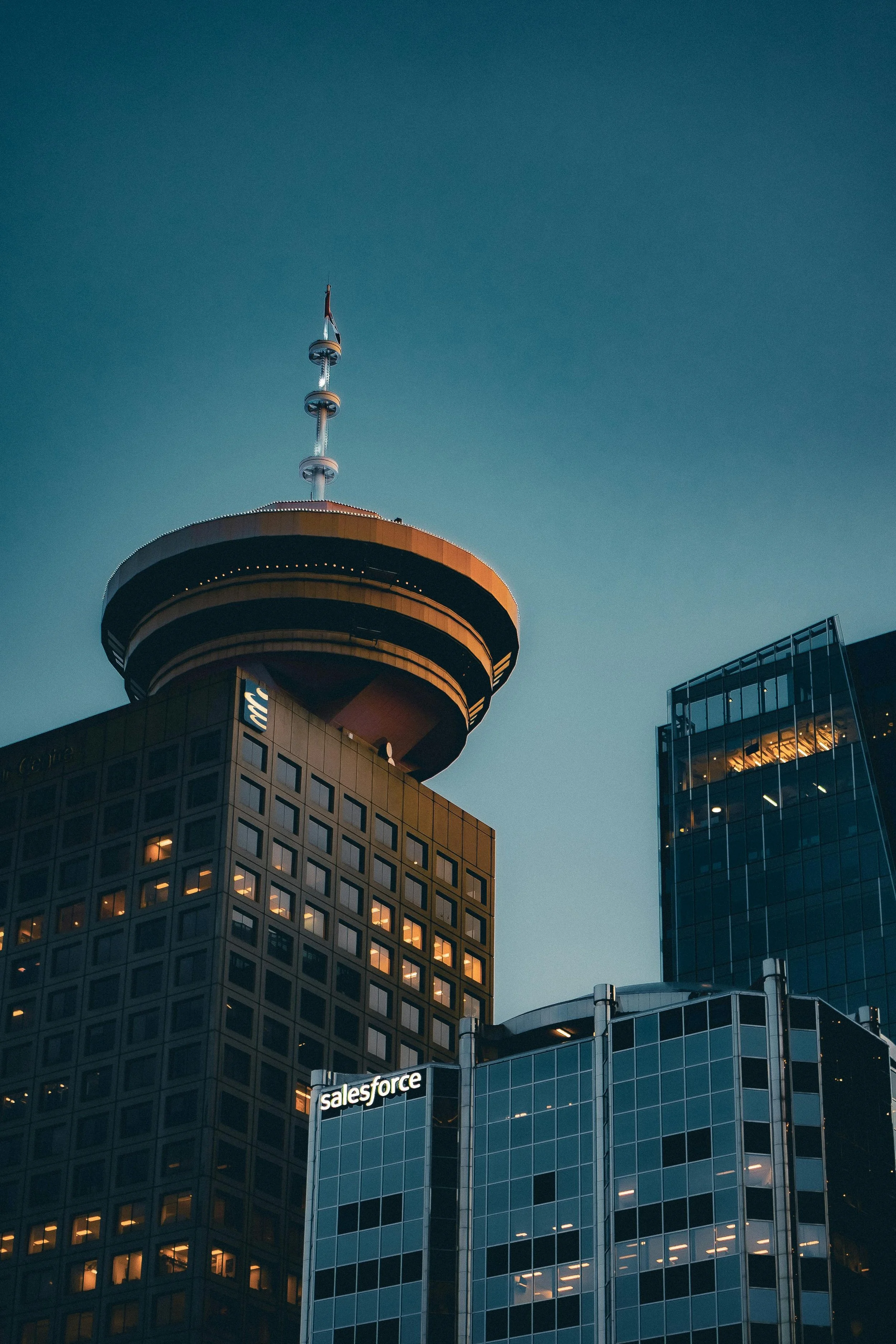 Skyscraper buildings in a city with illuminated windows and signs, including a prominent Salesforce sign, and a tower with a round observation deck near the top against a clear sky.