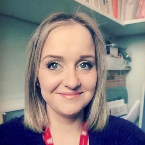 A woman with blonde hair and blue eyes smiling in an indoor setting with shelves and books in the background.