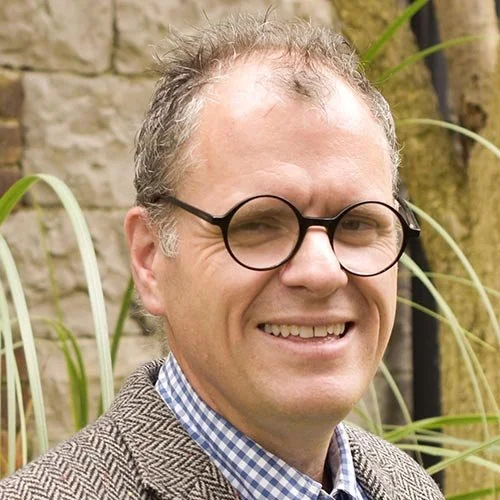 A man wearing black round glasses, a gray checkered blazer, and a blue and white checkered shirt, smiling outdoors with greenery and a stone wall in the background.