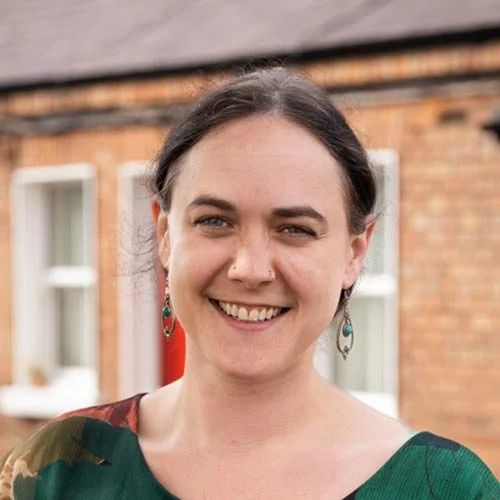A woman with dark hair smiling outdoors in front of a brick building with windows.