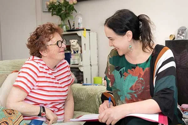 Two women are sitting and smiling at each other in a living room, having a conversation.