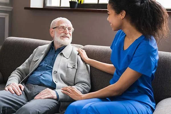 A nurse talking and smiling at an elderly man sitting on a sofa in a healthcare setting.