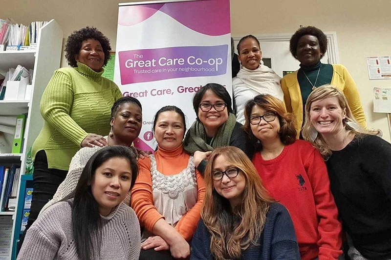 Group of ten women of diverse backgrounds smiling and posing together indoors, with a banner behind them that reads 'The Great Care Co-op.' The background includes shelves with binders and documents.
