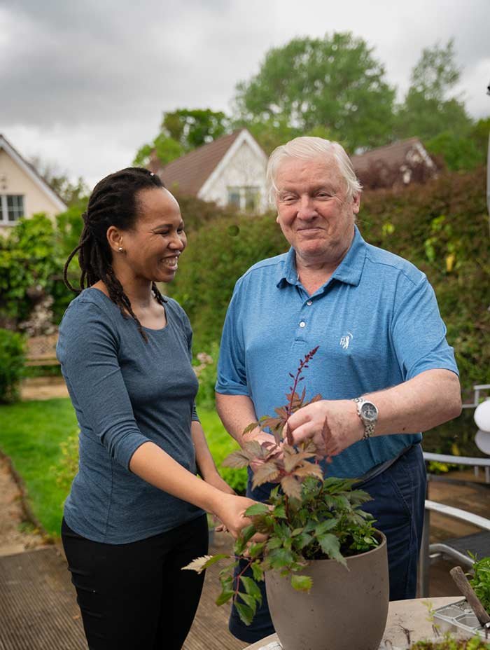 A woman and an older man smiling while gardening together outside in a backyard with greenery and houses in the background.