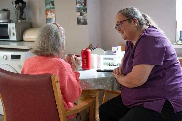 A caregiver and an elderly woman having a conversation at a dining table in a home kitchen.