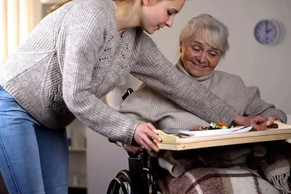 A young woman helping an elderly woman in a wheelchair to eat a meal at a table.