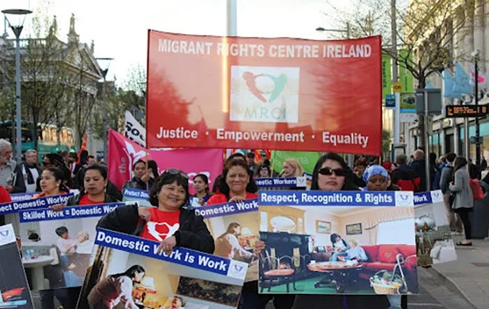 People holding signs and banners at a rally or march for domestic workers' rights. The signs promote respect, recognition, and the importance of domestic work, with a large red banner in the background from the Migrant Rights Centre Ireland.