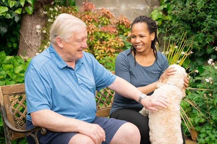 An elderly man and a young woman sitting on a garden bench, petting a small dog.