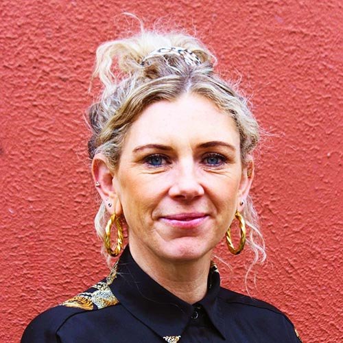 A woman with blonde curly hair in a bun, wearing large hoop earrings and a black shirt, standing in front of a textured red wall.