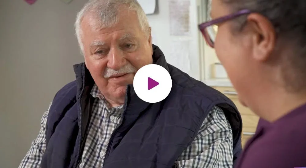 An elderly man and a woman are having a conversation, with the man looking at the woman and smiling. The woman is wearing glasses and facing him.