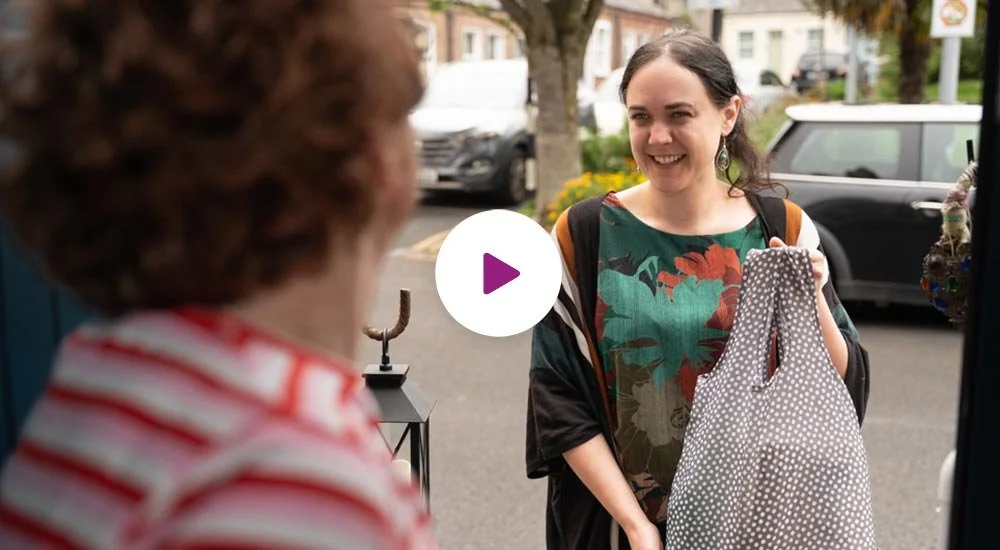 A young woman with dark hair smiling and holding a bag while talking to an elderly woman at a store entrance, with cars and trees in the background.