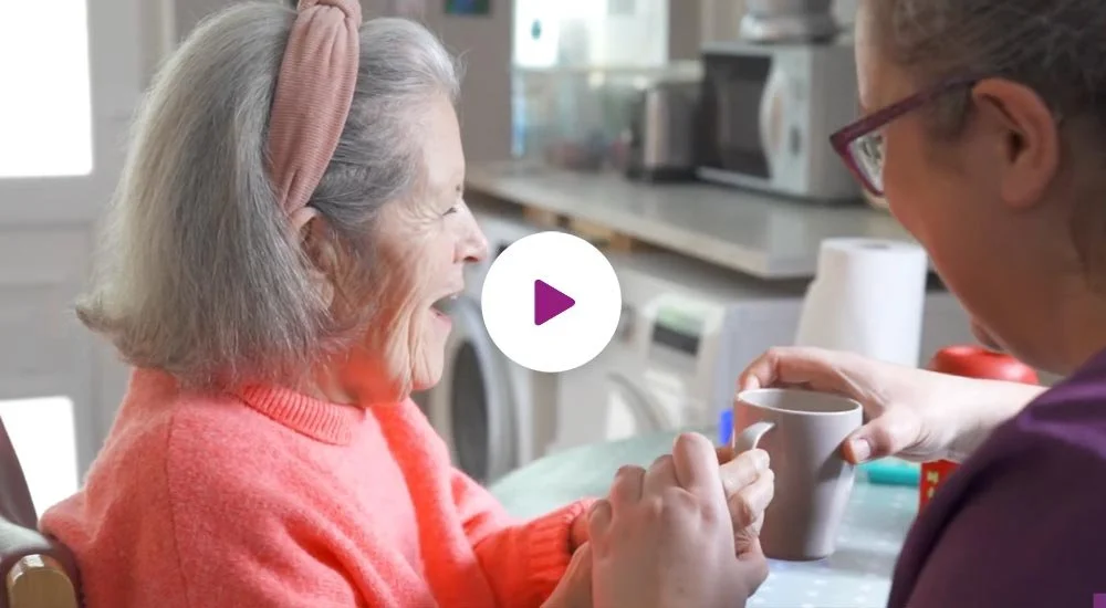 A young woman giving a cup to an elderly woman who is laughing and wearing a pink sweater and headband in a kitchen setting.