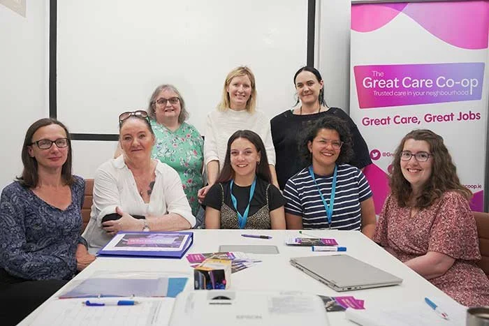 Group of seven women sitting and standing around a conference table at a meeting, with a pink banner in the background that reads 'The Great Care Co-op.'