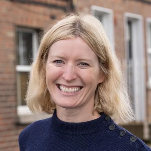 A smiling woman with blonde hair wearing a navy blue sweater, standing outdoors in front of a brick building with white window frames.
