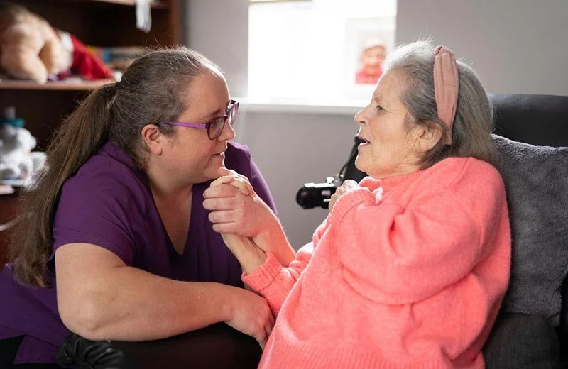 A young woman and an elderly woman are holding hands and looking at each other affectionately in a cozy room.