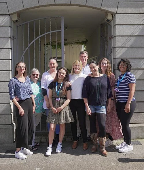Group of nine diverse women and men standing in front of a gated entrance to a building, smiling for the photo.
