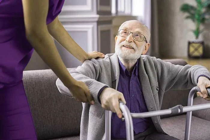 An elderly man with a walker smiling at a caregiver in a purple uniform who is assisting him in an indoor setting.
