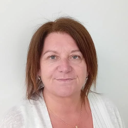A middle-aged woman with shoulder-length reddish-brown hair, wearing a white top and small earrings, standing against a plain light background.