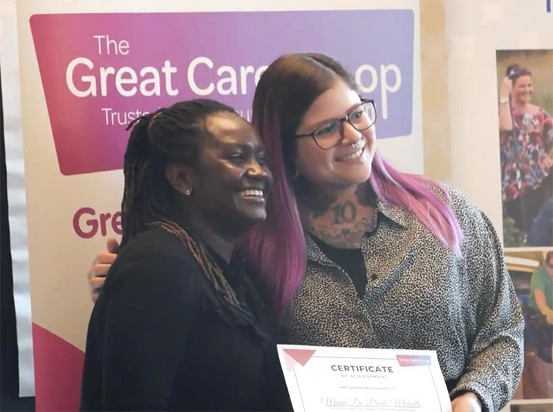 Two women smiling and holding a certificate at an event, standing in front of a pink and purple banner that reads 'The Great Card Shop'.