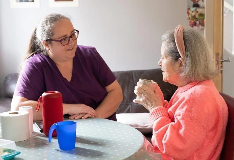 An elderly woman with a pink sweater and headband drinks from a mug while talking to a woman in a purple shirt at a table with colorful cups and paper towels.