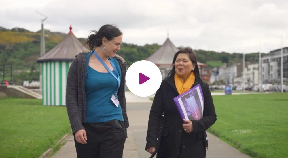 Two women walking outdoors on a path, engaged in conversation, with green grass, a striped tent-like structure, and buildings in the background.