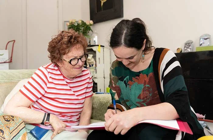 Two women sitting on a sofa, one older woman in a red and white striped shirt, and a younger woman with dark hair and a patterned dress, looking at a notebook.