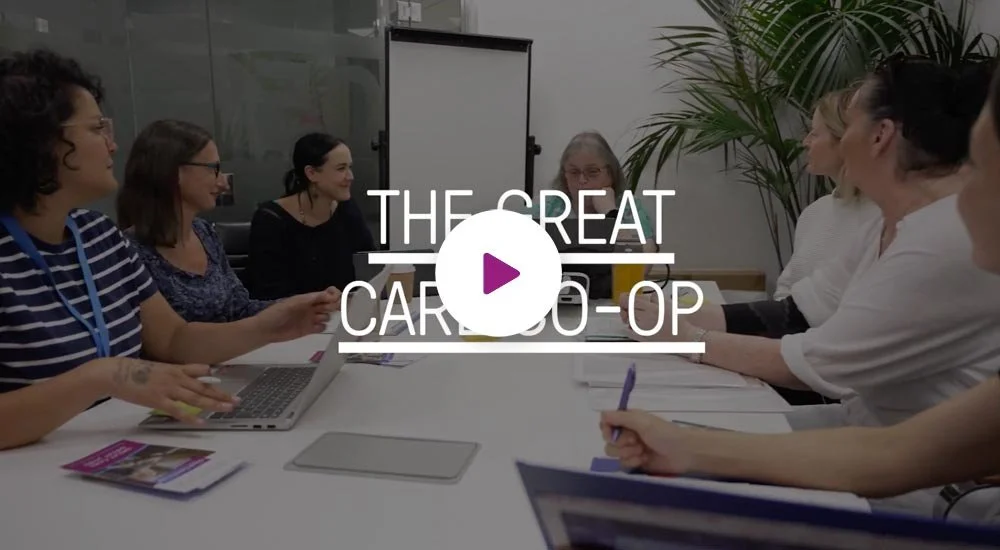 A group of women sitting around a conference table in a meeting room, engaged in discussion with laptops and papers. The screen in the background displays the title "THE GREAT CARDO-OP".