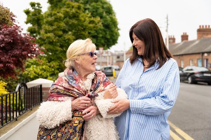 Two women smiling and engaging in conversation on a sidewalk with trees and houses in the background.