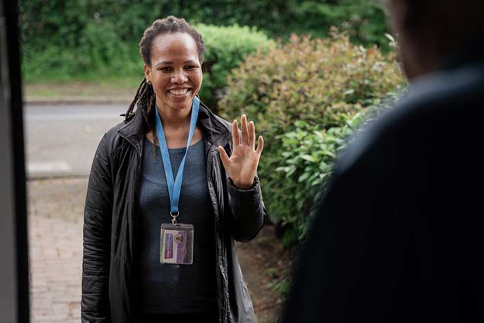 A smiling woman with a lanyard greeting an unseen person outdoors.