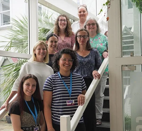 Group of nine women standing on indoor stairway, smiling, with plants and glass windows in background.