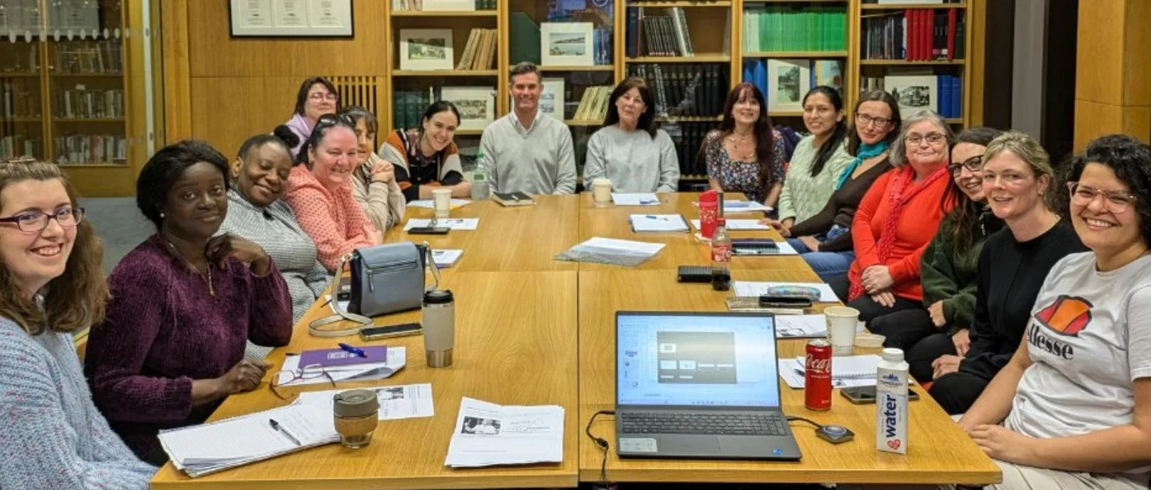 A group of 15 people sitting around a large wooden conference table in a library room, smiling at the camera. The table has laptops, notebooks, papers, drinks, and snacks on it.