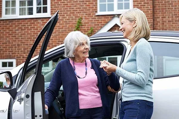 Two women, one elderly with gray hair and the other middle-aged with blonde hair, smiling and talking outside near a silver car with the driver's door open.