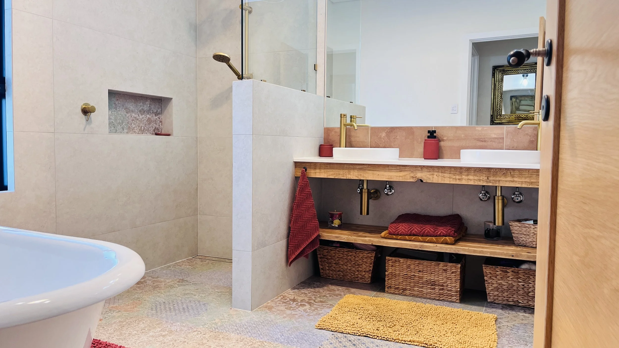 A bathroom with beige tiled walls and floor, a walk-in shower with glass enclosure, a double sink vanity with two brass faucets, a large mirror, and red accessories including soap dispensers and towels. Underneath the vanity are baskets and a yellow 